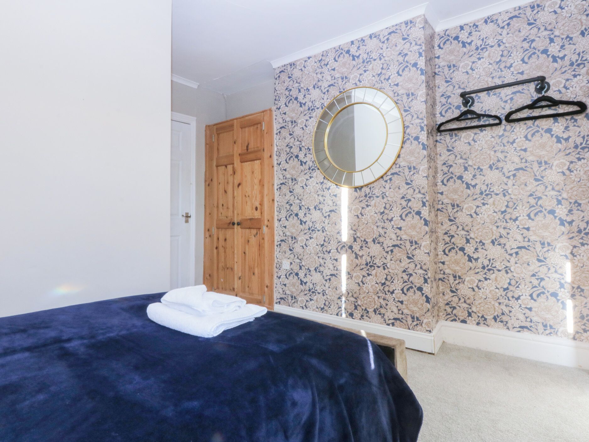A bedroom with a bed, a wooden wardrobe, a round mirror, and a wall with floral wallpaper.
