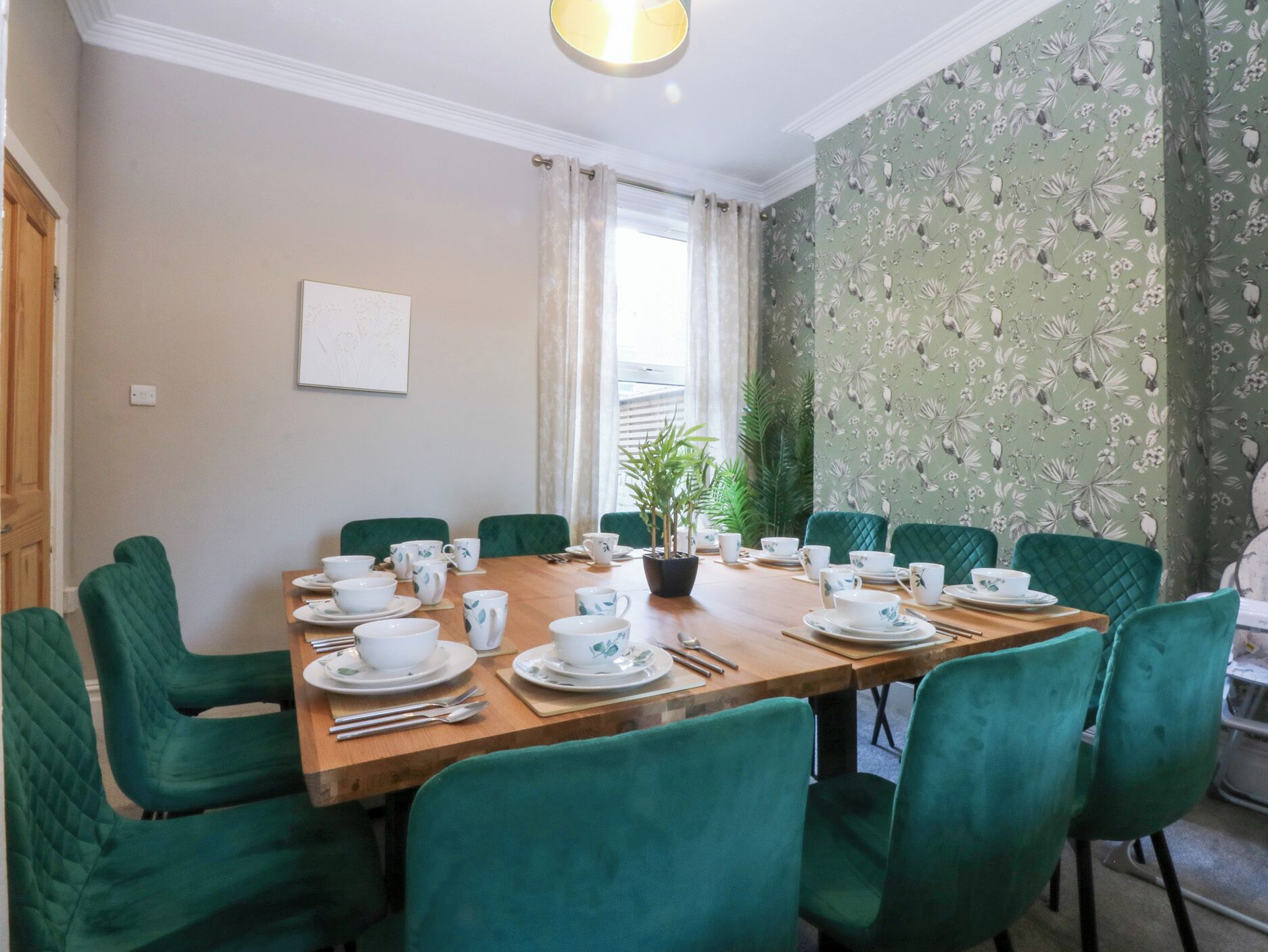 A dining area with a long wooden table set for a meal, surrounded by green upholstered chairs.