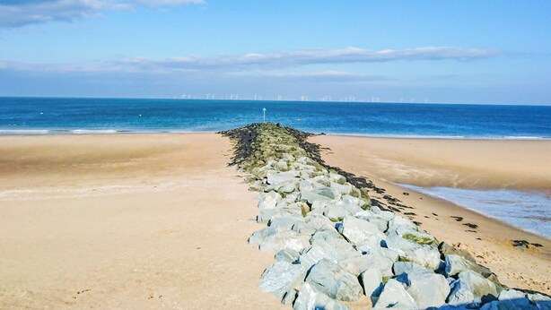 Una spiaggia con un molo frangiflutti roccioso che si estende verso il mare, cielo sereno e turbine eoliche in lontananza.