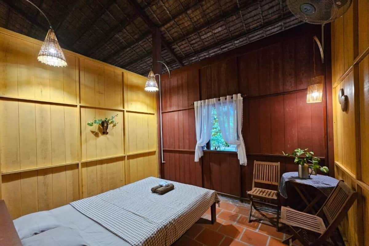 Ein Zimmer mit einem Bett, einem Tisch mit Stühlen, einem Ventilator und einem Fenster mit Vorhängen.