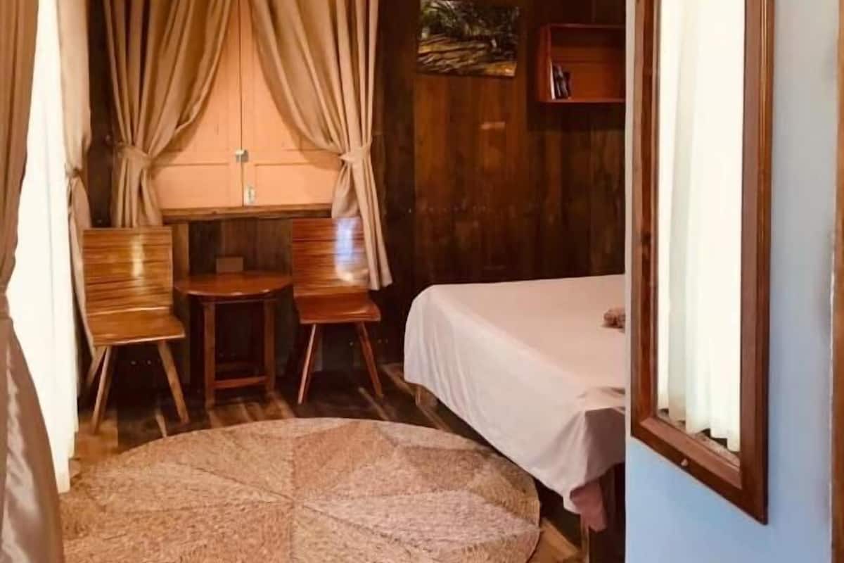 Ein Zimmer mit einem Bett, zwei Stühlen, einem kleinen Tisch, einem runden Teppich, einer Klimaanlage und einem Fenster mit Vorhängen.