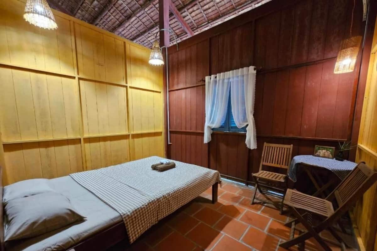 Ein Zimmer mit einem Bett, einem kleinen Tisch, zwei Stühlen, einem Fenster mit Vorhängen und einer Deckenleuchte.