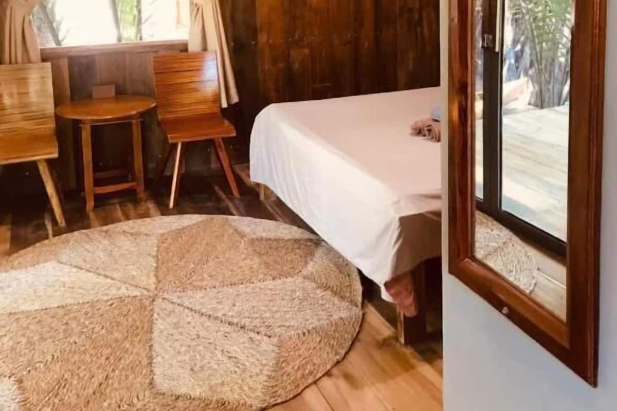 Ein gemütliches Zimmer mit einem Bett, einem kleinen Tisch und einem Stuhl. Es gibt ein Fenster mit Vorhängen, eine Wand-Klimaanlage und ein Regal mit einer Pflanze.