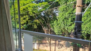 A view from a window with a metal railing, a concrete ledge, and a street with a parked car and trees in the background. - Studio in Butantã, near USP. (São Paulo)