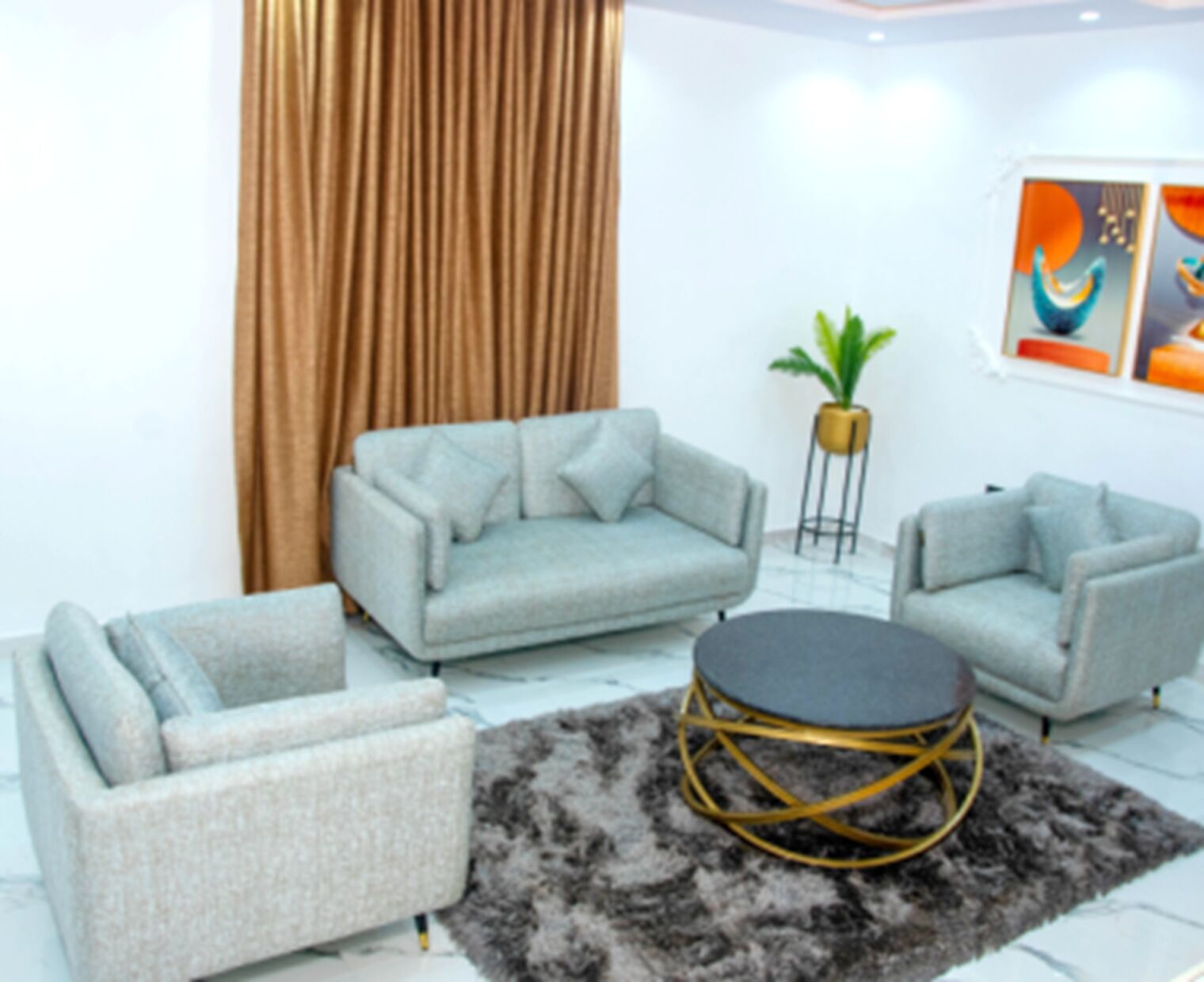 A modern living room with a grey sofa, a matching armchair, and a round coffee table with a gold frame. A large curtain and a potted plant are also visible.