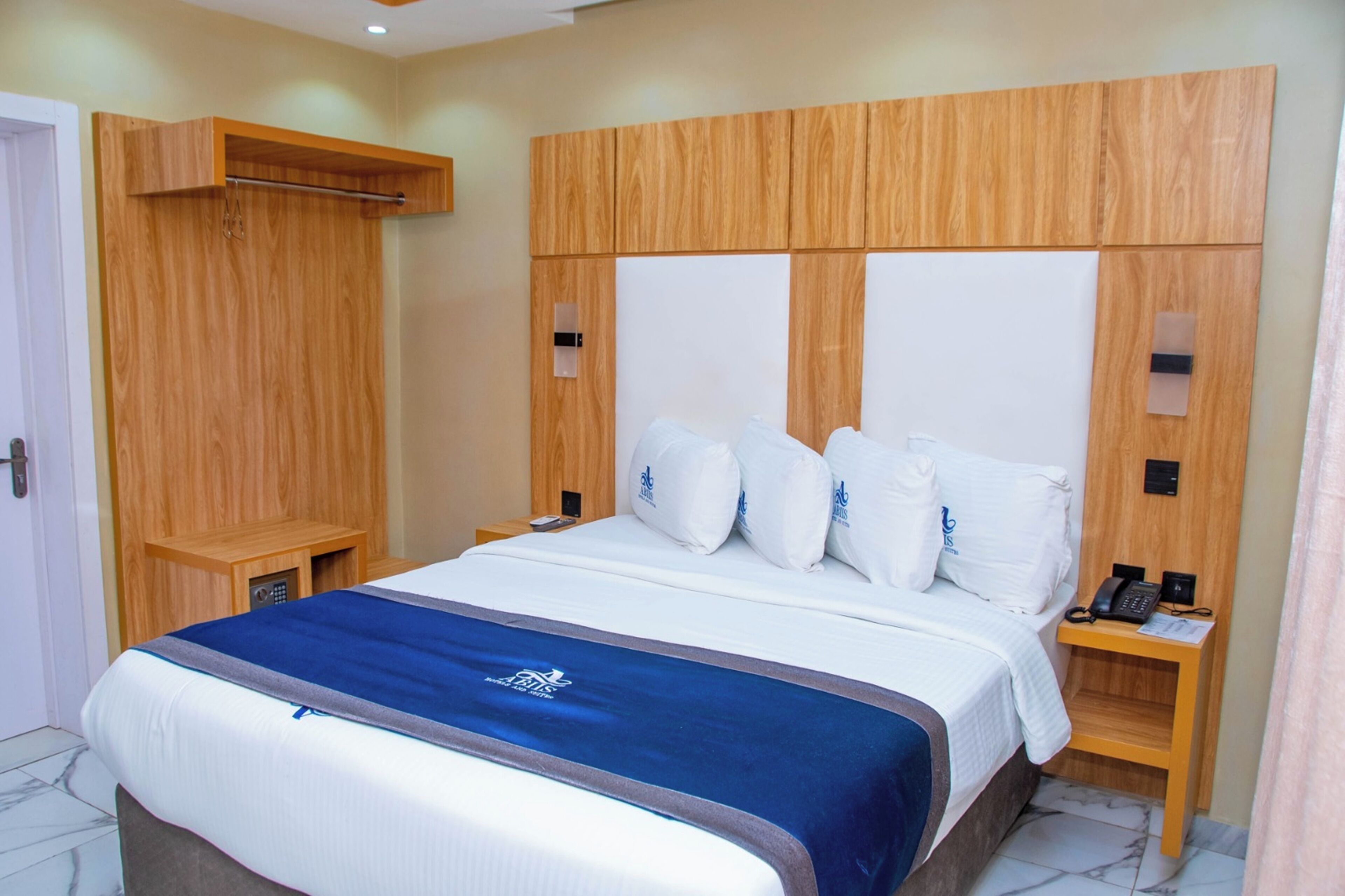 A hotel room with a wooden headboard, a bed with white bedding and a blue accent, a bedside table with a phone, and a small shelf.