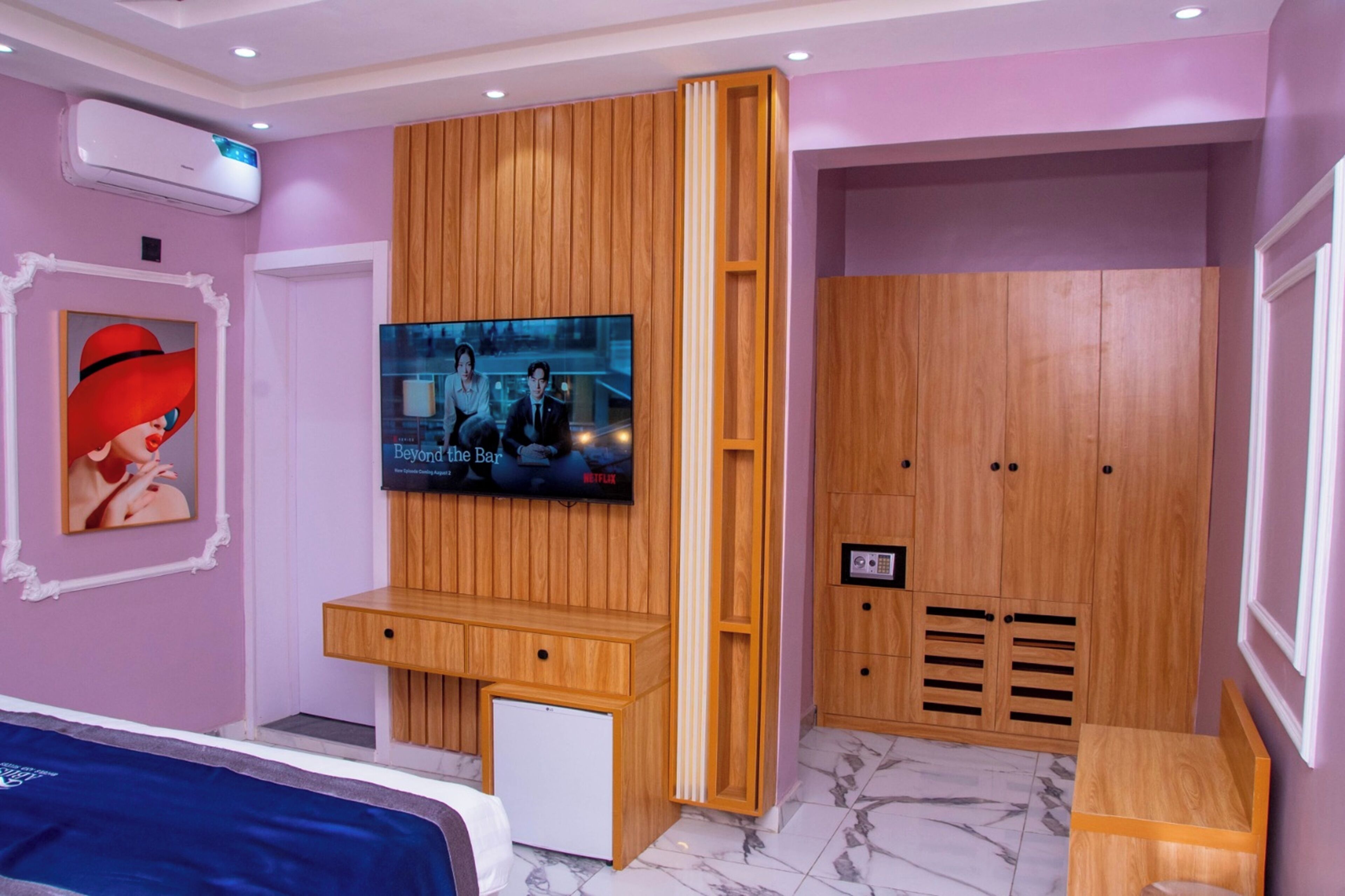 A modern bedroom with a TV mounted on a wooden panel wall, a small desk, a wardrobe, and a bed with a blue bedspread.