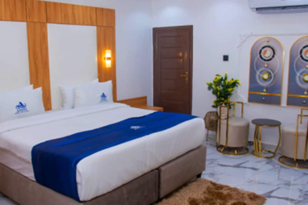 Une chambre d’hôtel avec un grand lit, une tête de lit en bois, un tapis marron, une petite table ronde et une plante en pot.