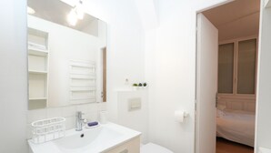 Ein modernes Badezimmer mit Waschbecken, Spiegel und Toilette. Ein kleines Fenster mit Jalousien und eine Tür zum Schlafzimmer.