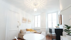 Ein heller, weißer Raum mit einem runden Tisch und Stühlen, einem Sofa mit gelber Decke und einem Kamin.