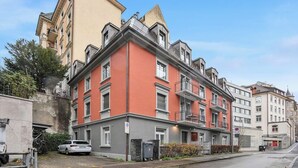 A multi-story building with a red facade, balconies, and a parking area in front. - Premium studio in city center - Color 3 (Zürich)