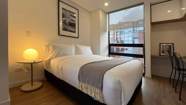 Une chambre à coucher moderne avec un grand lit, une table de chevet avec une lampe, un tableau encadré au mur et une vue sur les bâtiments par la fenêtre.