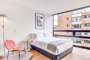 A modern bedroom with a large bed, a red chair, a floor lamp, and a view of a brick building through a large window. - w Beautiful Loft in the Heart of Bogot (Bogotá)