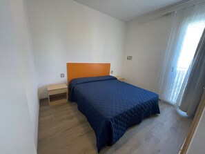A bedroom with a bed, a nightstand, a wooden floor, and a window with curtains. - Sunny Family Apartment Steps From the Beach (San Michele al Tagliamento)