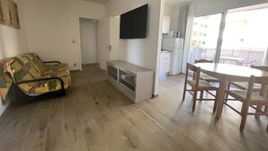 A compact living space with a sofa, a TV mounted on the wall, a dining table with chairs, and a kitchenette in the background. - Sunny Family Apartment Steps From the Beach (San Michele al Tagliamento)