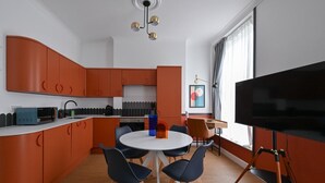 A modern kitchen with orange cabinets, a white dining table with chairs, a flat-screen TV on a stand, and a window with curtains. - Stylish 1-bed Apartment Near Angel Station (London)