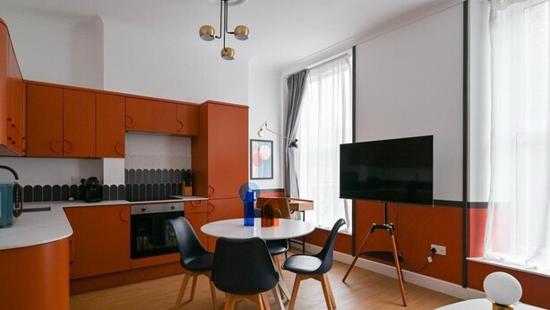 A modern kitchen with orange cabinets, a dining area with black chairs, and a large TV on the wall. - Stylish 1-bed Apartment Near Angel Station (London)