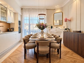 Een eetkamer met een ronde tafel voor zes personen, een keuken met moderne apparatuur en een groot raam met dunne gordijnen.