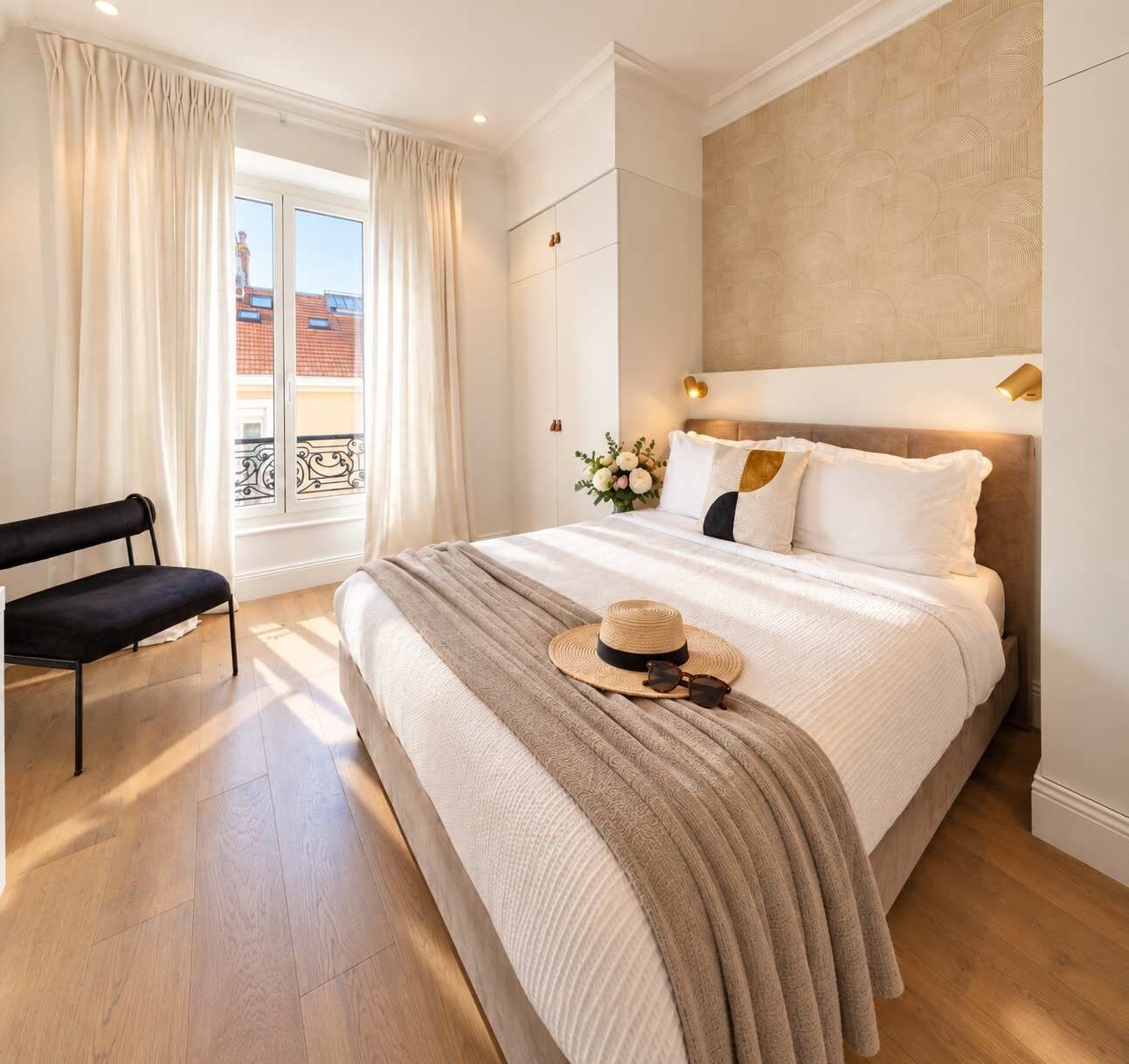 A bedroom with a large bed, a bench, a window with curtains, and a view of buildings outside.