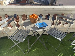 Ein runder Tisch mit zwei Metallstühlen, einem Korbwäscher mit Orangen und einem blauen Buch darauf.