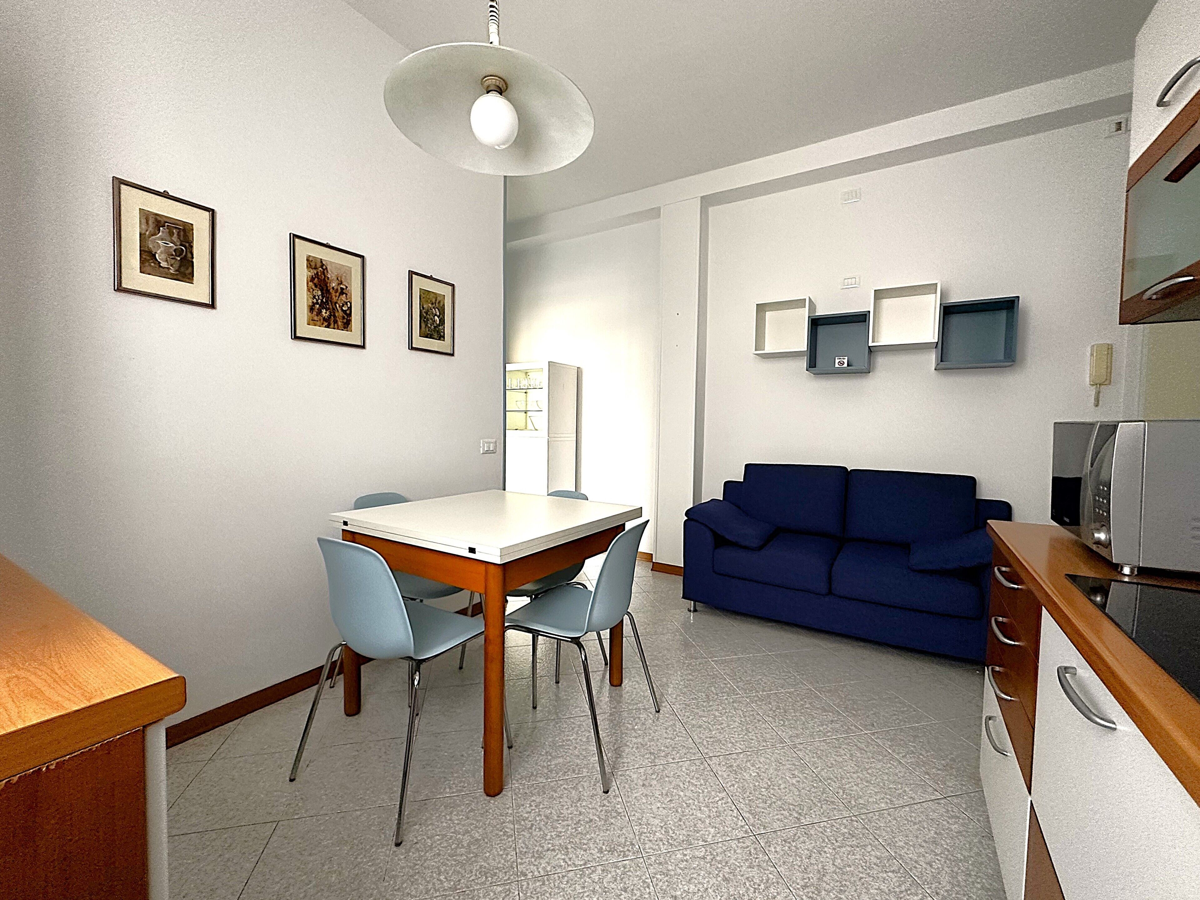 A compact living space with a dining table and chairs, a blue sofa, a kitchenette with a microwave, and framed pictures on the wall.