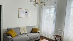 Ett vardagsrum med en grå soffa, två gula kuddar, ett träbord, en inramad tavla på väggen och en kristallkrona.