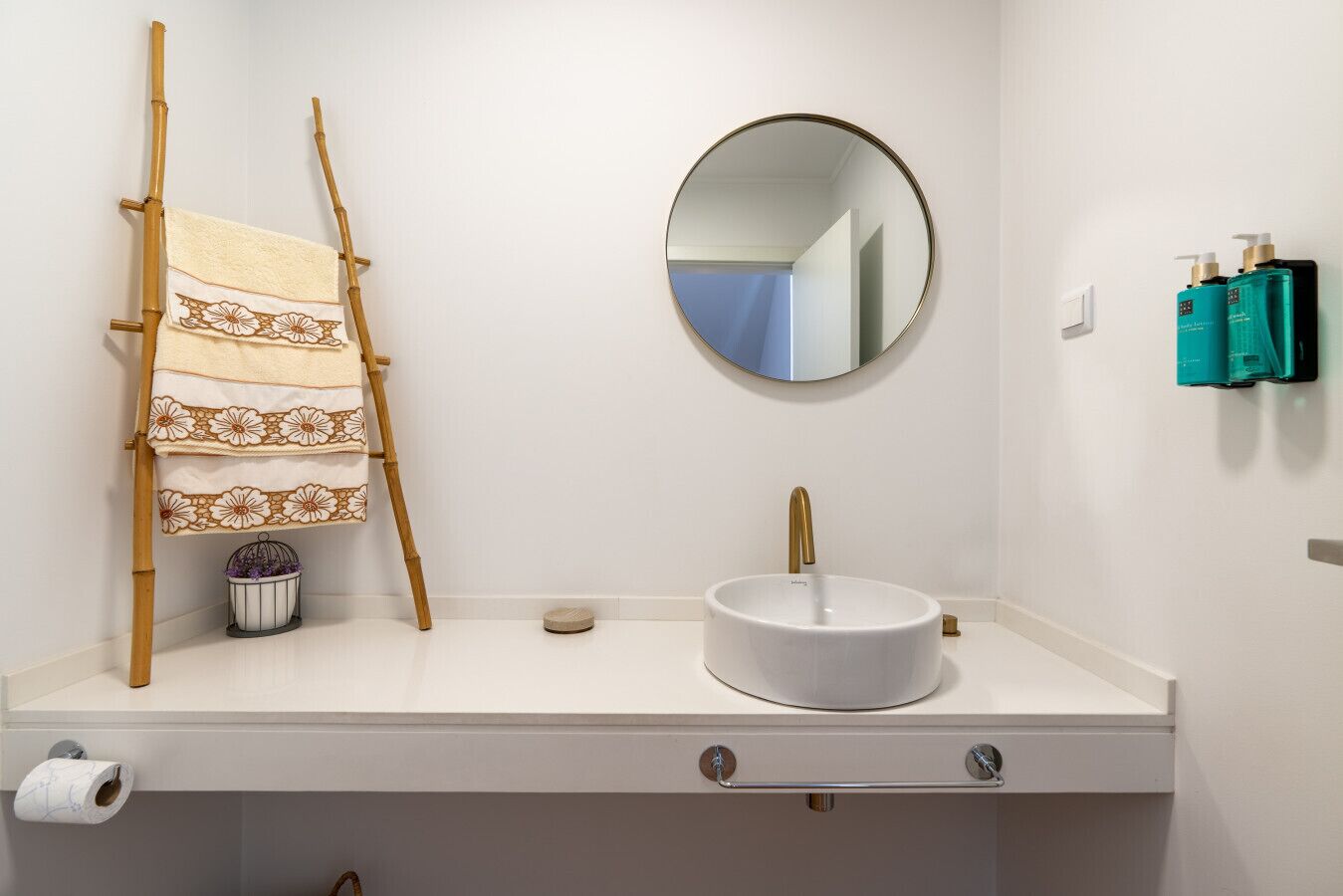 Une salle de bain avec un lavabo blanc, un miroir rond, un porte-serviettes avec des serviettes à motifs et deux flacons de produits de toilette accrochés au mur.