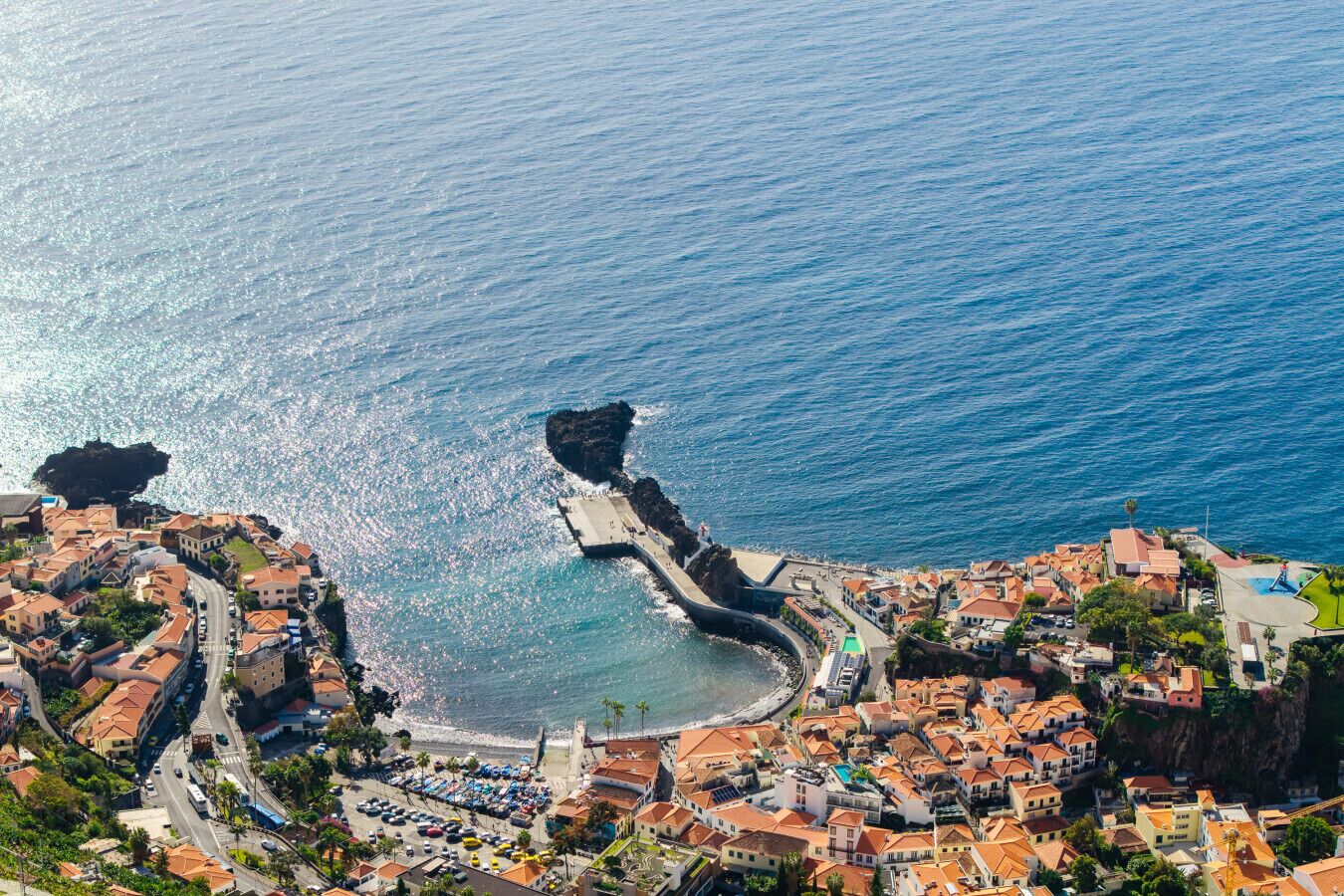 An aerial view of a coastal town with a prominent rocky coastline, a marina, and a mix of residential buildings with red-tiled roofs.