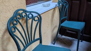 Deux chaises métalliques de couleur turquoise, dotées de dossiers décoratifs, sont placées sur un sol en béton, à côté d’un mur beige et d’une porte en bois.
