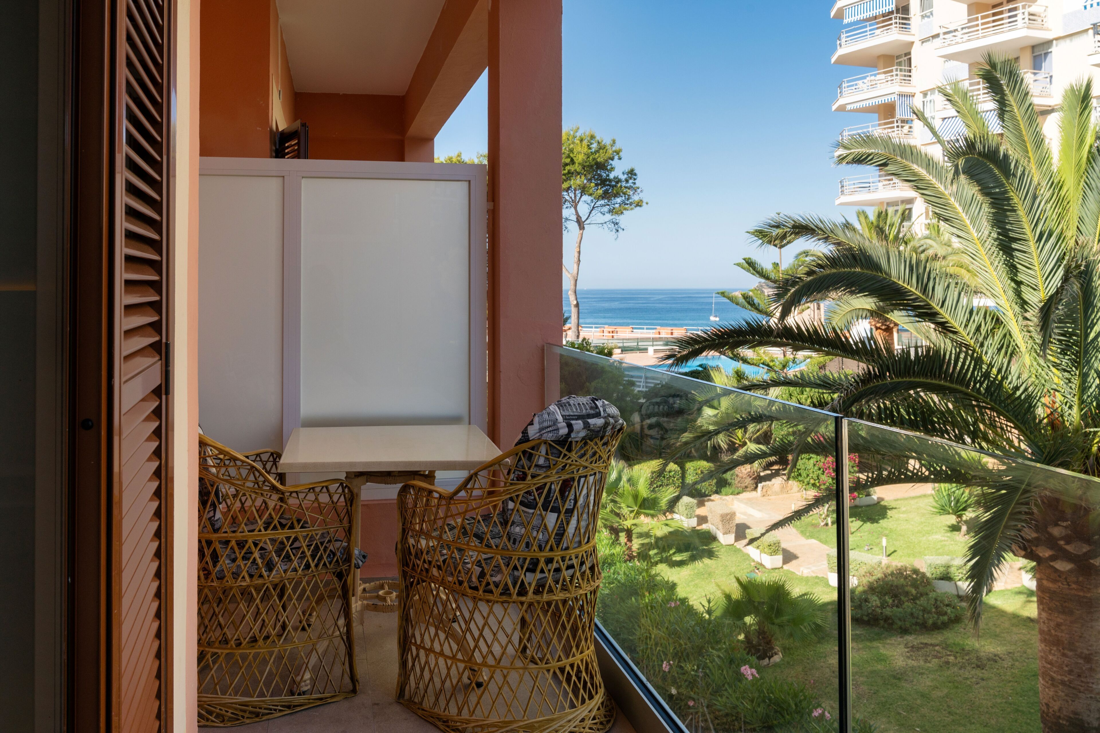 A balcony with wicker chairs, a white table, and a view of the sea and greenery.