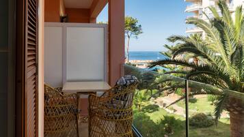 A balcony with wicker chairs, a white table, and a view of the sea and greenery.