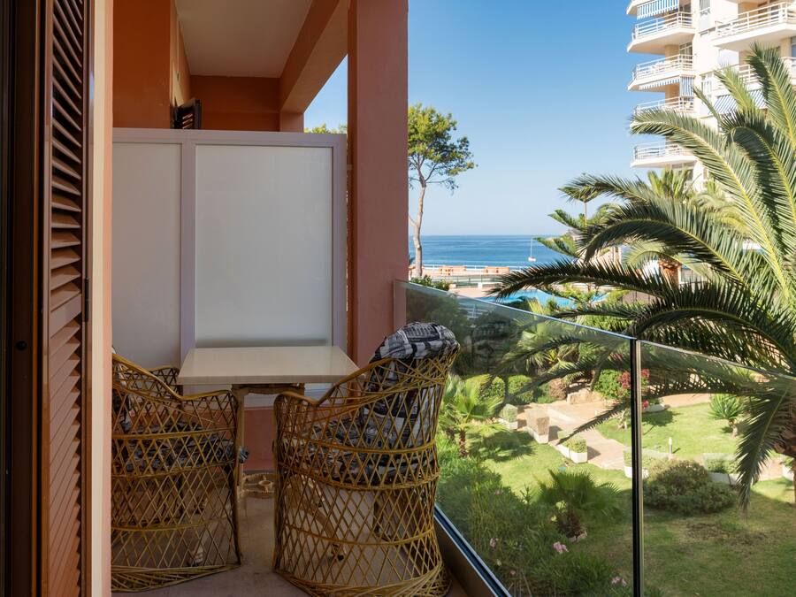 A balcony with wicker chairs, a white table, and a view of the sea and greenery.
