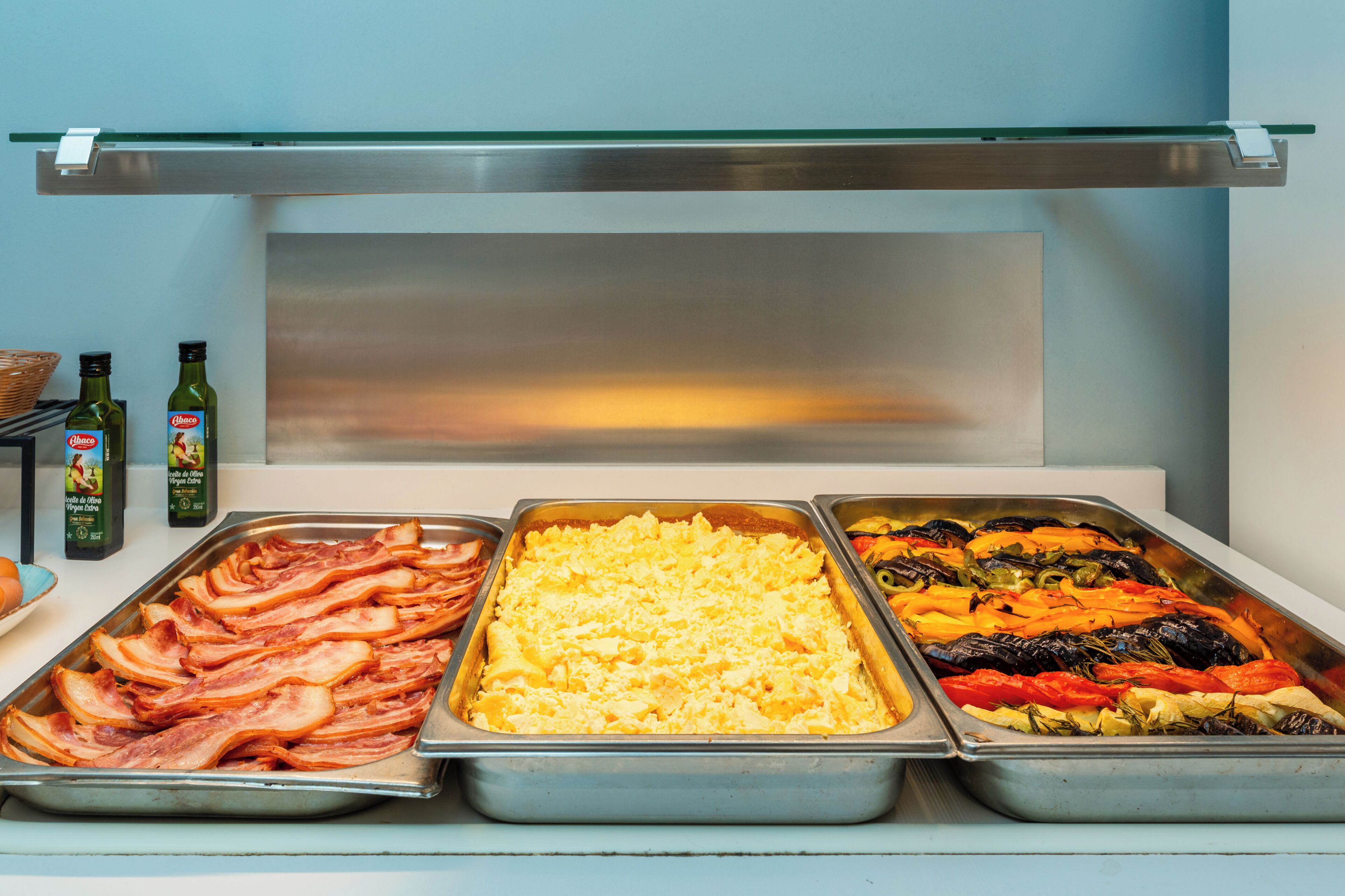 Buffet com diversas opções de comida, incluindo bacon, ovos mexidos e legumes assados.