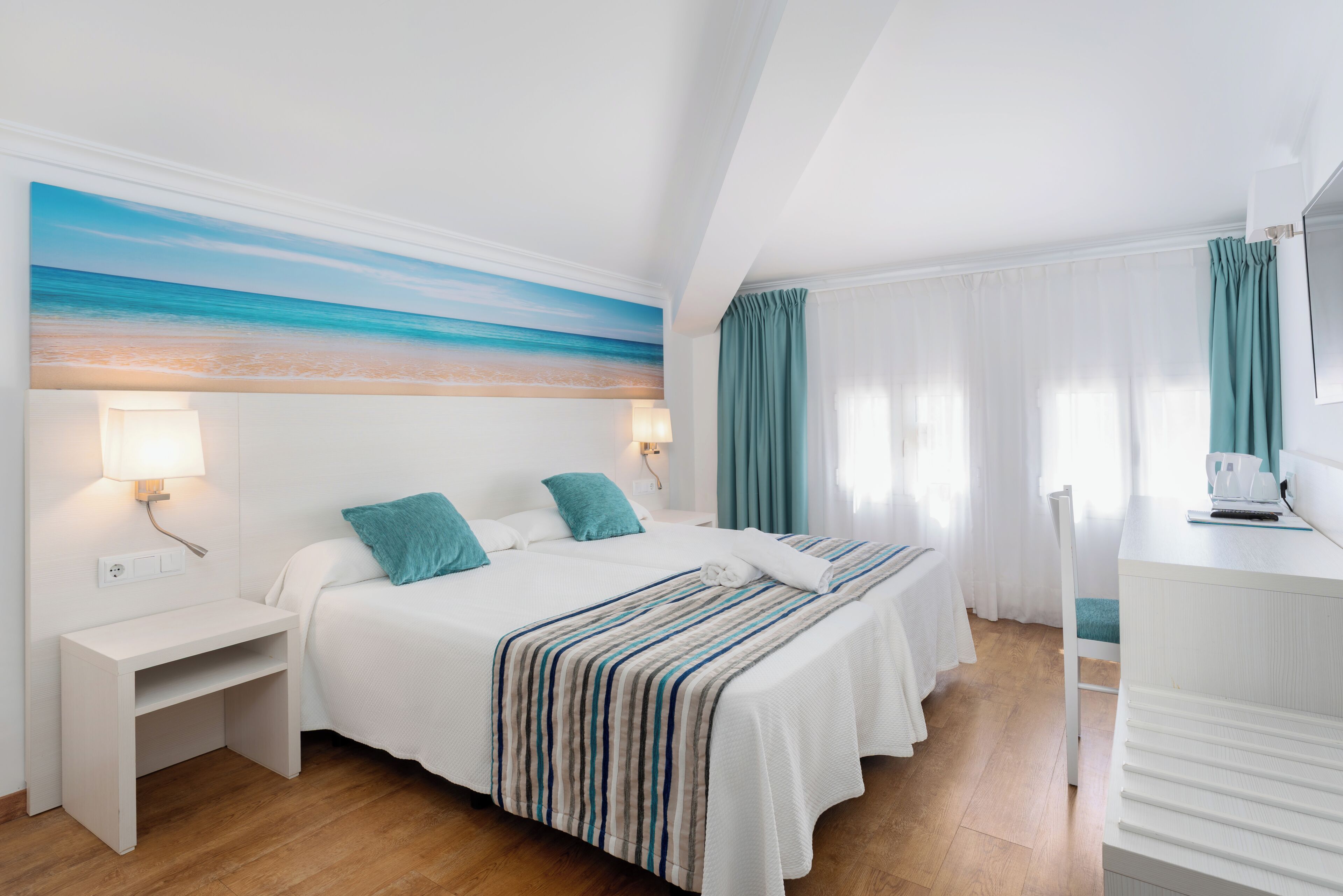 A hotel room with a bed, bedside tables, a desk, a chair, a desk lamp, a painting of a beach, and a window with curtains.