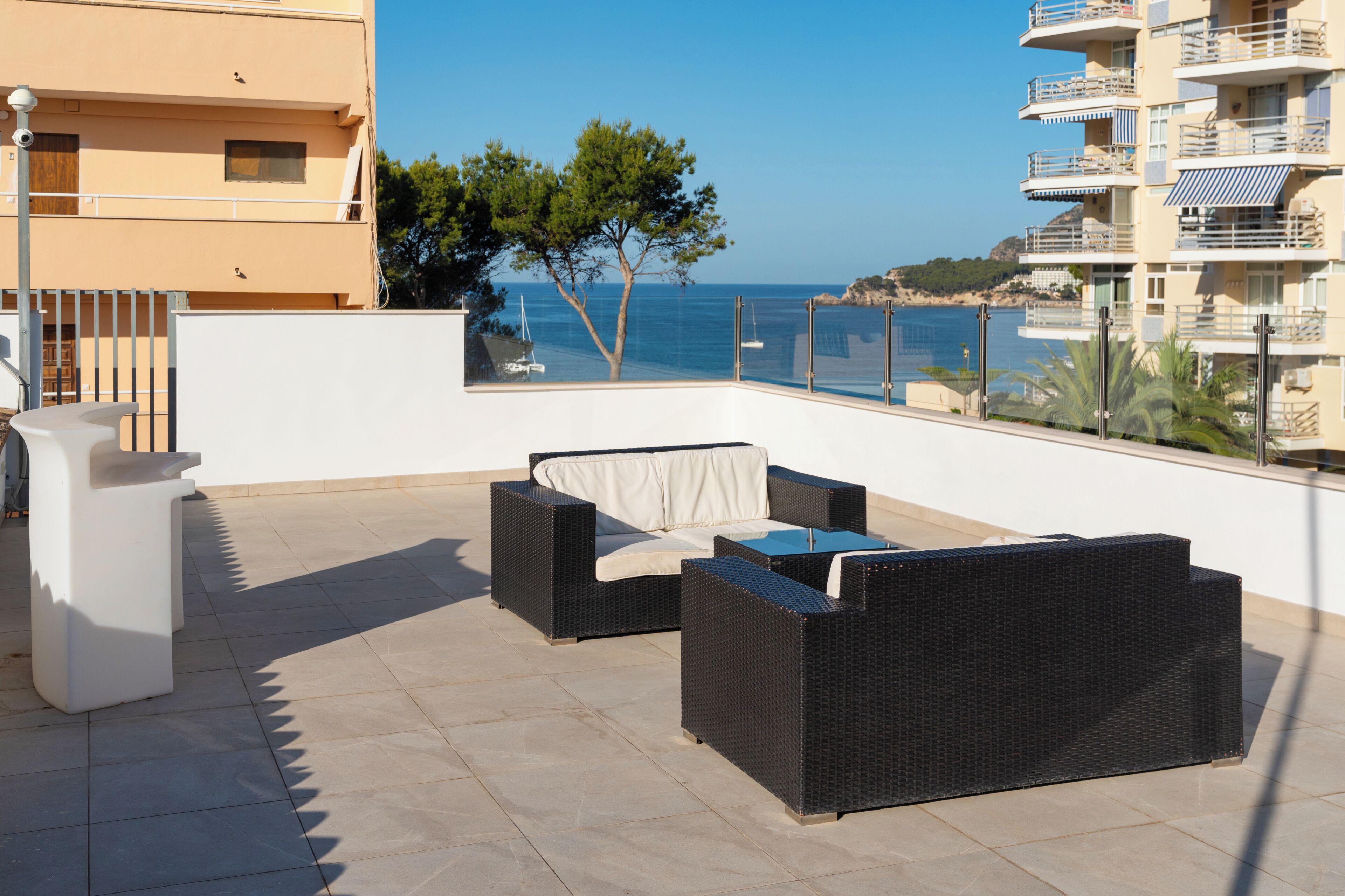 A balcony with wicker furniture, a white column, and a view of the sea and buildings.