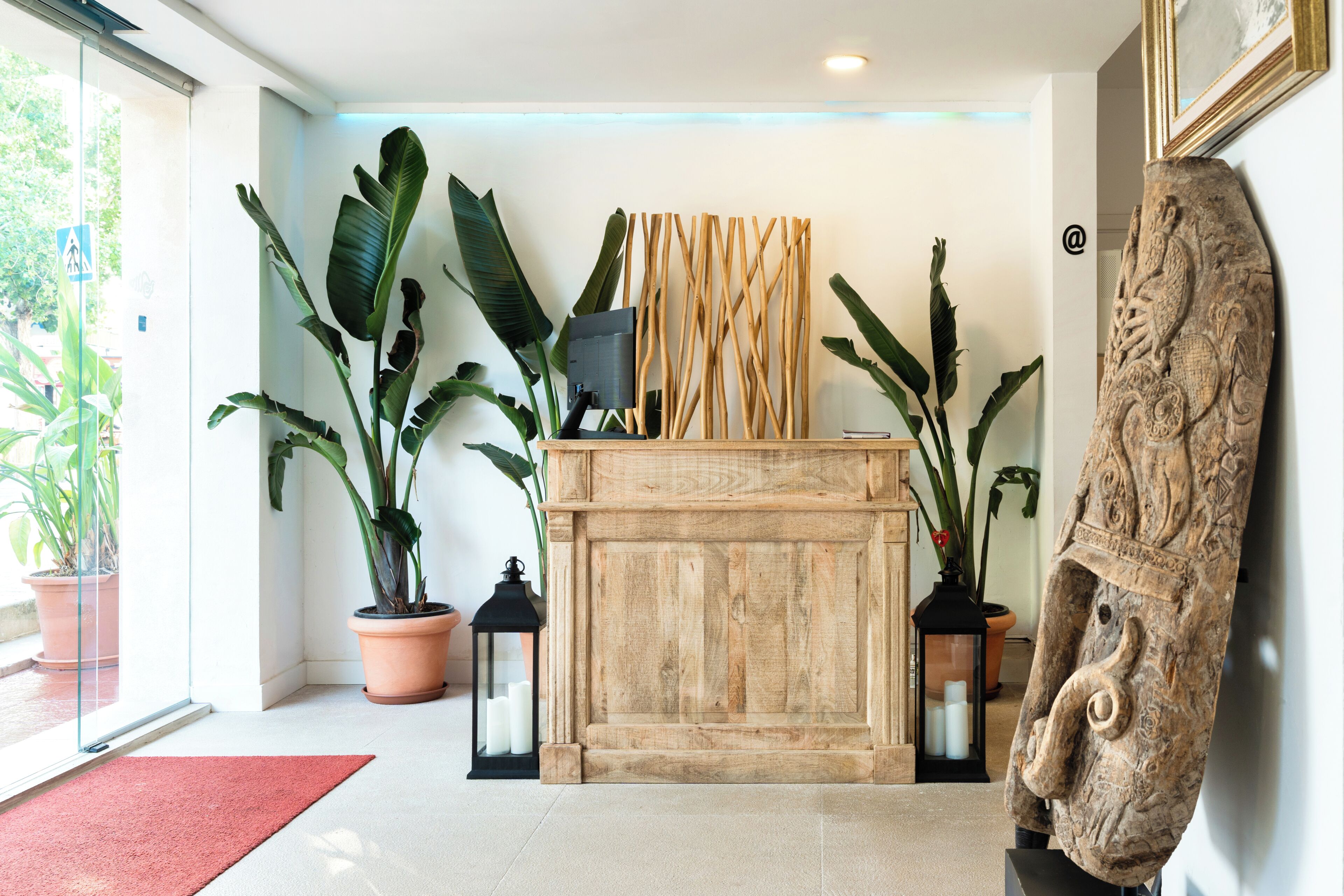 Uma área de recepção com uma mesa de madeira, plantas em vasos, uma escultura na parede e uma porta de vidro que dá para o exterior.