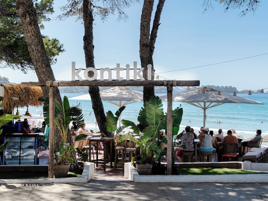 A beachside restaurant with outdoor seating, a thatched roof, and a view of the sea.