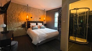 A bedroom with a stone wall, a bed with white bedding, two bedside lamps, a desk with a lamp, and a glass-enclosed shower area. - Hôtel Le Sakura  (Morestel)