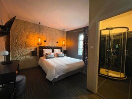Quarto com parede de pedra, cama com roupa de cama branca, duas luminárias de cabeceira, escrivaninha com abajur e área de chuveiro com proteção de vidro.