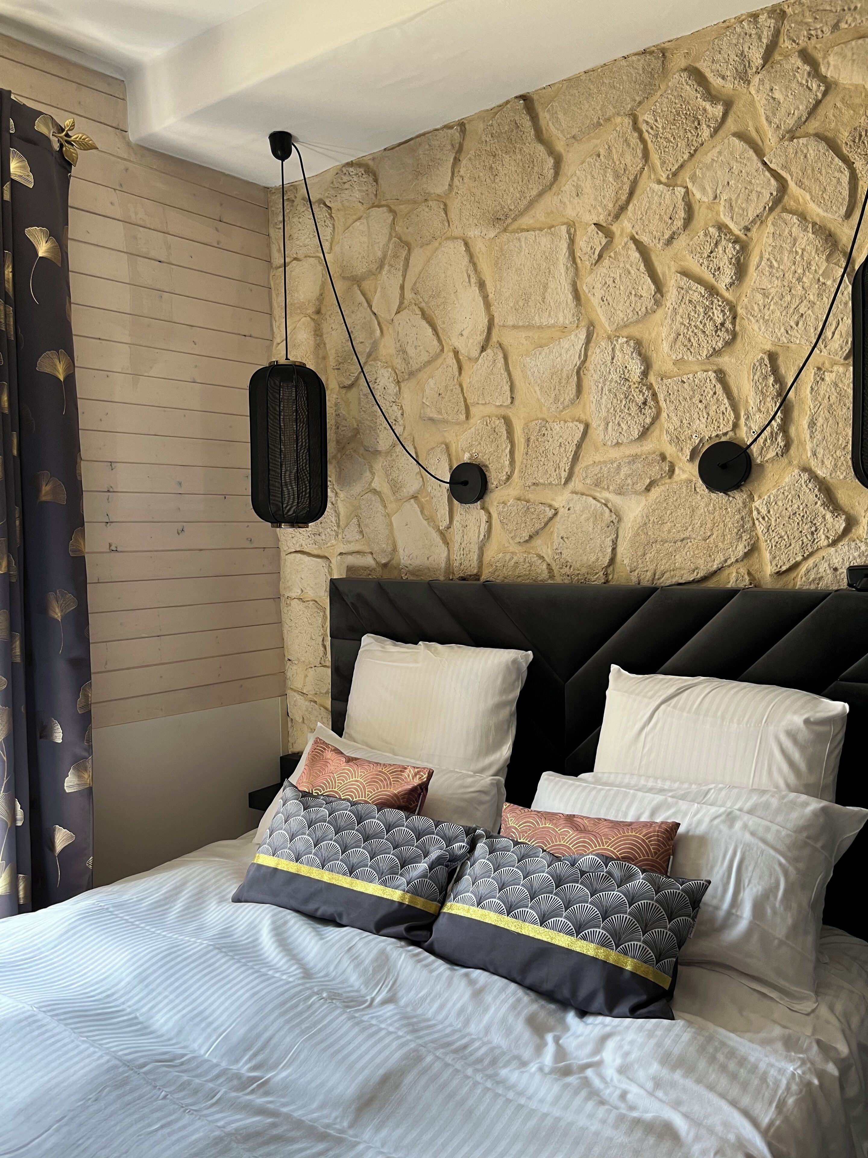 Un dormitorio con pared de piedra, una cama con muchos almohadones y dos lámparas colgantes.