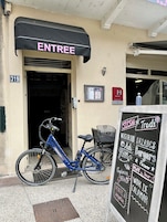 Fachada de loja com um letreiro que diz "ENTREE" e um quadro-negro com menu anunciando sushi e saladas. Uma bicicleta azul está estacionada na frente.