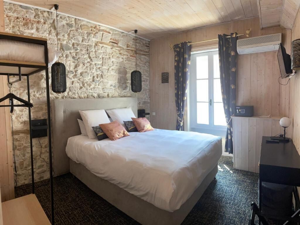 Een slaapkamer met een stenen muur, een bed met wit beddengoed, een bureau met een computer en een raam met gordijnen.