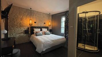 Een slaapkamer met een stenen muur, een bed met meerdere kussens, een bureau met een lamp en een douchecabine met glazen wanden.