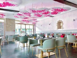 Um restaurante com mesas e cadeiras redondas, decorações florais cor-de-rosa no teto e vista para o exterior através de grandes janelas.