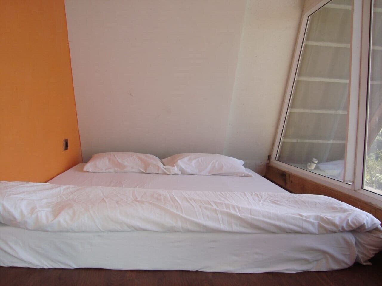 Un lit simple avec une literie blanche, dans une pièce dont un mur est orange et qui possède une fenêtre.