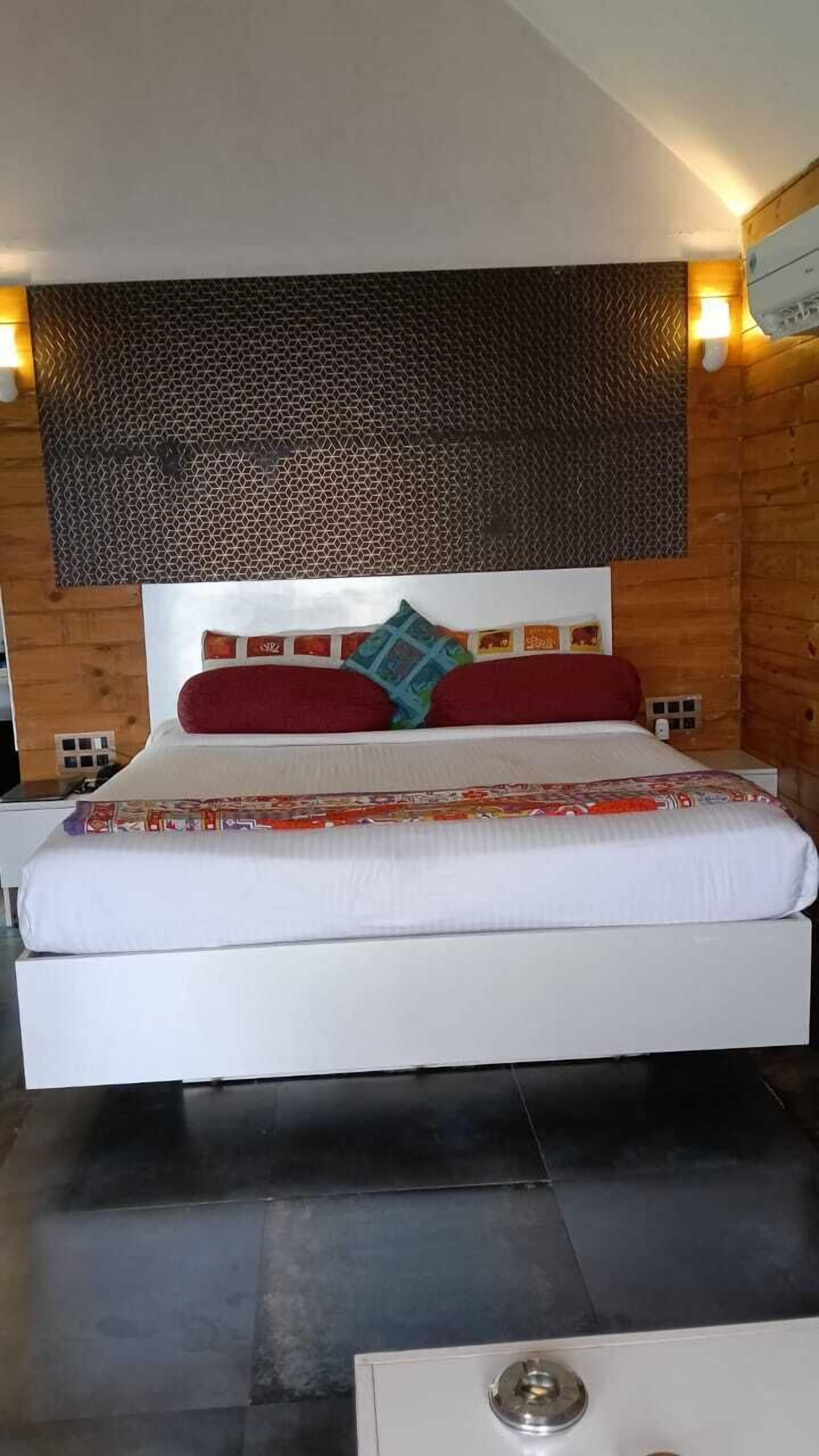 A bed with a white mattress and red and blue pillows, a patterned headboard, and a small bedside table with a lamp.