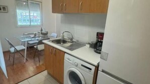 Une cuisine de taille réduite, dotée de meubles en bois, d’un lave-linge, d’un évier et d’une fenêtre avec des stores.