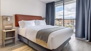 Een moderne hotelkamer met een groot bed, een nachtkastje, een wandlamp en een uitzicht op de stad door grote ramen.