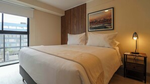 Un lit bien fait, recouvert d’une couverture beige, une table de chevet avec une lampe et un tableau encadré au mur.