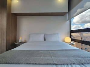 Quarto de hotel moderno com uma cama grande, mesa de cabeceira, abajur e janela com vista para a cidade.